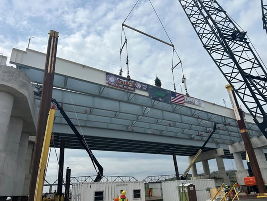Construction crew placing the final girder into place.  Crane is lowering it into place.  Various flags are on the side of the girder in celebration.